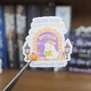 K&ouml;nnte beinhalten: Ein gestanzter Aufkleber mit einer violetten, gew&ouml;lbten Bibliothekst&uuml;r und einem wei&szlig;en Geist, der ein gr&uuml;nes Buch liest. Der Text lautet WELCOME TO THE HAUNTED READING CLUB und Stay until the last chapter. Er enth&auml;lt einen K&uuml;rbis und zwei Laternen.