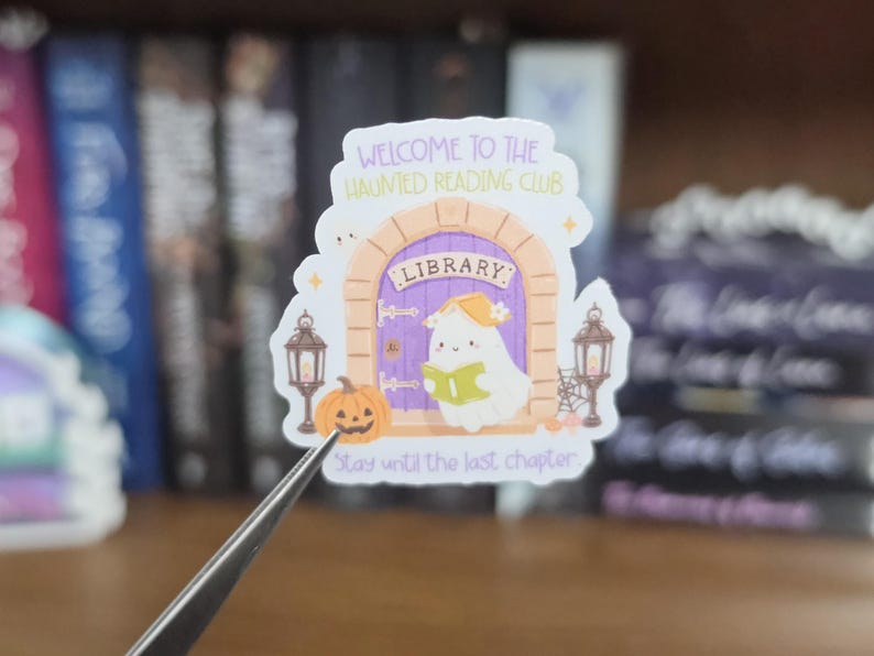K&ouml;nnte beinhalten: Ein gestanzter Aufkleber mit einer violetten, gew&ouml;lbten Bibliothekst&uuml;r und einem wei&szlig;en Geist, der ein gr&uuml;nes Buch liest. Der Text lautet WELCOME TO THE HAUNTED READING CLUB und Stay until the last chapter. Er enth&auml;lt einen K&uuml;rbis und zwei Laternen.