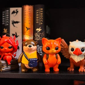 May include: Four colorful figurines of fantasy creatures on a shelf. A red figure with flame-like hair, a brown otter with a yellow shirt and blue bag, an orange cat with a backpack, and a white and orange griffin with wings.