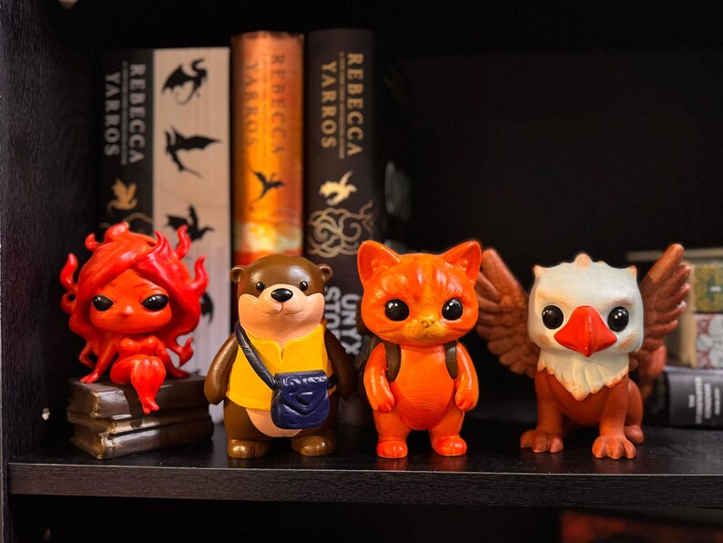 May include: Four colorful figurines of fantasy creatures on a shelf. A red figure with flame-like hair, a brown otter with a yellow shirt and blue bag, an orange cat with a backpack, and a white and orange griffin with wings.