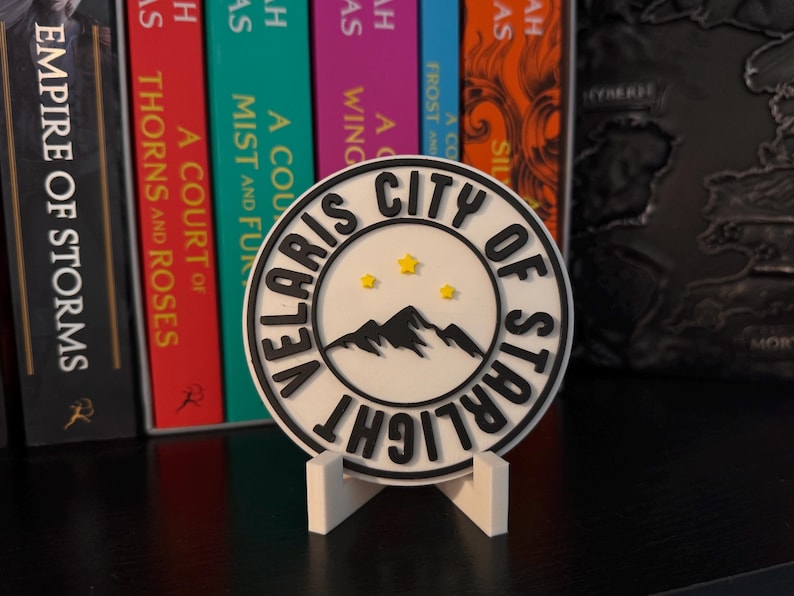 May include: A white and black circular coaster with the text "VELARIS CITY OF STARLIGHT" and a mountain and star design. The coaster is displayed on a white stand, with colorful books in the background.