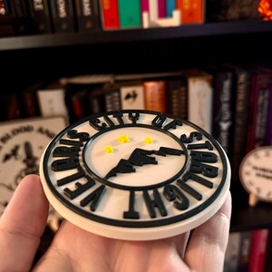 May include: A round, off-white coaster with black lettering and a black mountain silhouette. The text reads "VELVET CITY OF STARLIGHT". Three small yellow stars are in the center. The coaster is held in a hand.