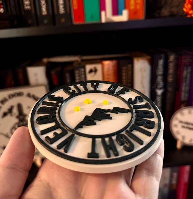 May include: A round, off-white coaster with black lettering and a black mountain silhouette. The text reads "VELVET CITY OF STARLIGHT". Three small yellow stars are in the center. The coaster is held in a hand.