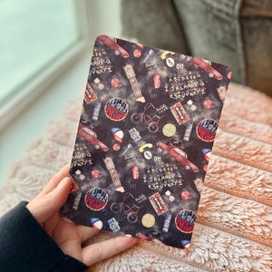 May include: A rectangular, patterned item featuring a dark background with various illustrations. The design includes a red car, a grandfather clock, a bicycle, and the number "011". The alphabet is also present. The item is held by a person.