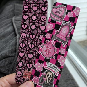 May include: Two rectangular bookmarks with pink and black designs. One features a repeating pattern of skulls, hearts, and flowers. The other has a pink and black checkerboard background with various stickers, including a throne, a ghost mask, and text.