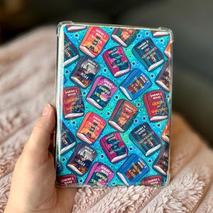 May include: A clear protective case for an e-reader, featuring a repeating pattern of book covers with titles like "Throne of Glass" and "Queen of Shadows" by Sarah J. Maas, set against a teal background with stars.