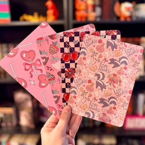 May include: A hand holds four pink decorative cards with various patterns. One card features a heart-shaped lollipop, another has cherries on a checkered background, and the others have skunks, hearts, and the text "Love Stinks".