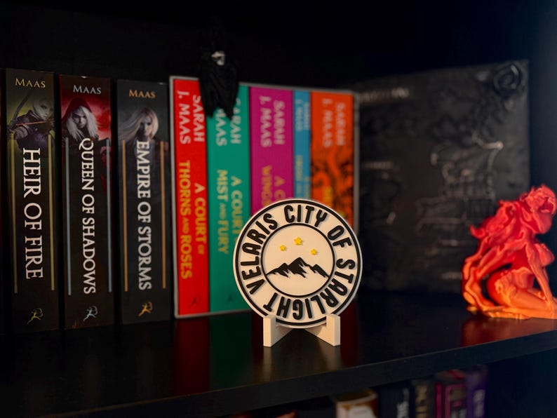 May include: A bookshelf displays a collection of books with titles like "Heir of Fire" and "Queen of Shadows." A round "Velaris City of Starlight" sign stands in front of the books. An orange sculpture is on the right side of the shelf.