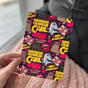 May include: A rectangular card with a colorful design featuring the text "DUNGEON CRAWLER CARL" in yellow and red. The card includes illustrations of a dinosaur, donuts, and other graphic elements on a dark background.