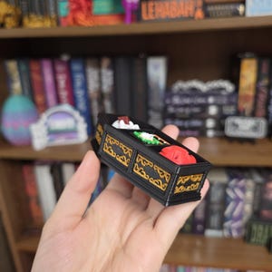 May include: A small, rectangular black box with gold-colored decorative filigree patterns along the sides. The top of the box is open, revealing three distinct compartments containing small, colorful sculpted items in white, green, and red.