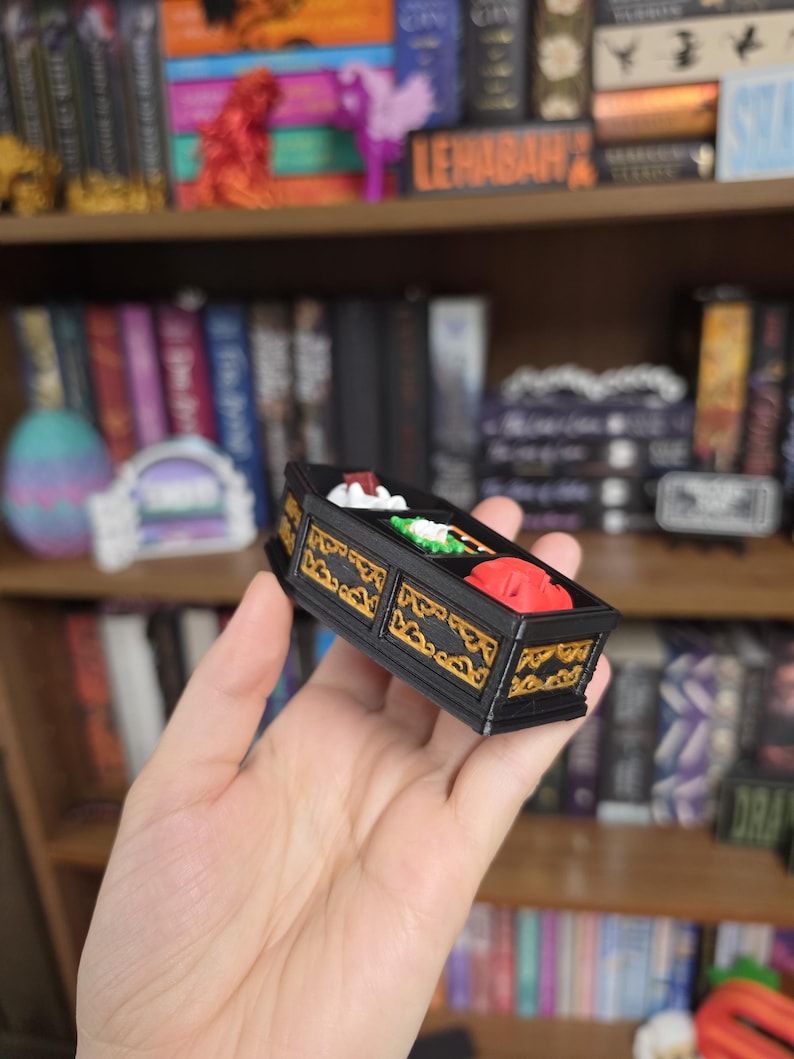 May include: A small, rectangular black box with gold-colored decorative filigree patterns along the sides. The top of the box is open, revealing three distinct compartments containing small, colorful sculpted items in white, green, and red.