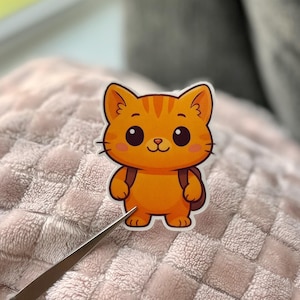 May include: A sticker featuring a cartoon orange cat with a backpack. The cat has large, round eyes, a small nose, and a smiling mouth. The cat's fur is orange with darker orange stripes on its head. The sticker is on a pink, textured surface.