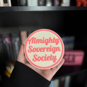 May include: A white round coaster with a pink border and the text "Almighty Sovereign Society" in pink lettering.