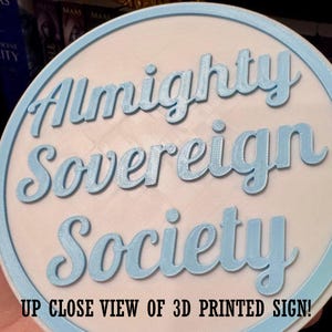 May include: A round, 3D-printed sign with the words "Almighty Sovereign Society" in light blue script. The sign has a light blue border and a white background. The text "UP CLOSE VIEW OF 3D PRINTED SIGN!" is at the bottom.