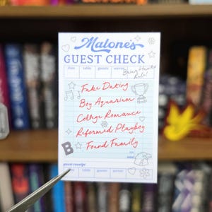 May include: A rectangular white guest check notepad with blue grid lines and blue text reading Malone's GUEST CHECK. Handwritten red text lists Fake Dating, Boy Aquarium, College Romance, Reformed Playboy, and Found Family with small icons.