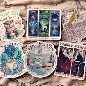 May include: A collection of colorful stickers featuring fantasy-themed designs. One sticker depicts an otter with mail, another a deer in a forest, and others with text like "Suriel Tea Co" and "Once Upon a Time".