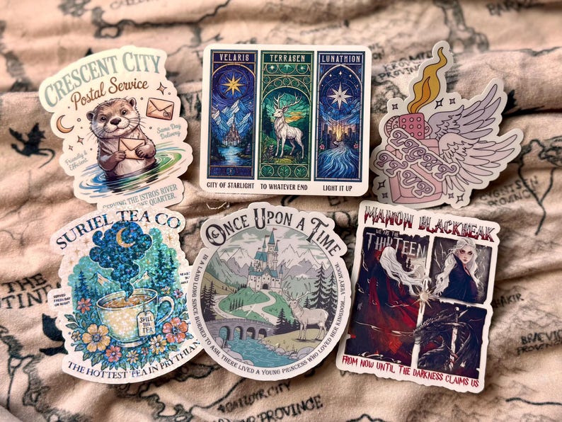 May include: A collection of colorful stickers featuring fantasy-themed designs. One sticker depicts an otter with mail, another a deer in a forest, and others with text like "Suriel Tea Co" and "Once Upon a Time".