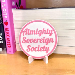 May include: A pink and white round sign with the text "Almighty Sovereign Society" in pink lettering. The sign is on a white stand.