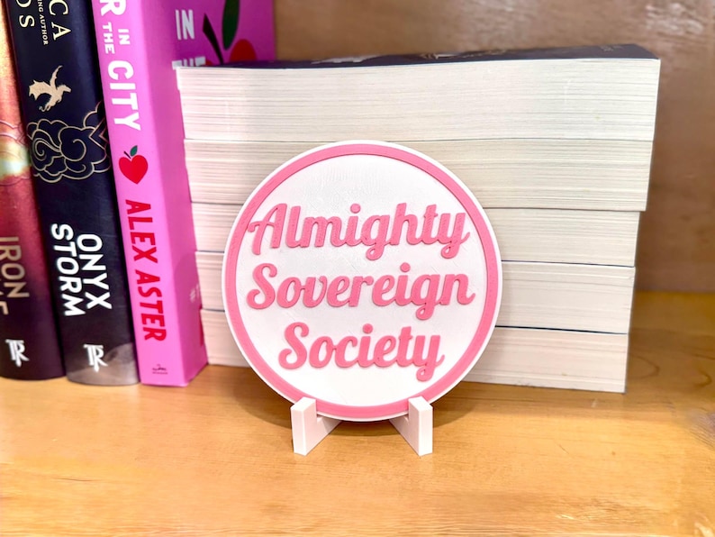 May include: A pink and white round sign with the text "Almighty Sovereign Society" in pink lettering. The sign is on a white stand.
