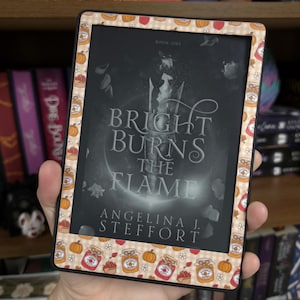 May include: An e-reader device featuring a decorative skin with a beige gingham pattern, orange pumpkins, and jars of preserves. The screen displays the book cover for Bright Burns the Flame by Angelina J. Steffort with a dark, moody illustration.