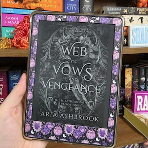 May include: An e-reader device featuring a protective case with a repeating pattern of purple and black Halloween-themed icons, including pumpkins and cats. The screen displays the book cover for Web of Vows and Vengeance by Aria Ashbrook.