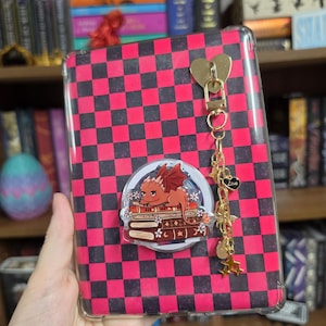 May include: A rectangular tablet case featuring a black and hot pink checkerboard pattern. An acrylic charm of a small red dragon resting on a stack of books is attached to the center. A gold-toned metal chain with heart and star charms hangs down.