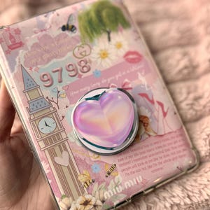 May include: A pink and clear phone case with a heart-shaped pop socket. The case features illustrations of a clock tower, bees, flowers, and text. The pop socket has a gradient pink and purple heart design.