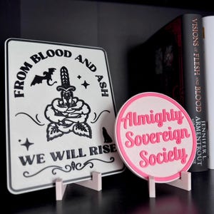 May include: Two white signs with black text and graphics. The sign on the left says "From Blood and Ash We Will Rise" with a black rose, a dagger, a bat, and a wolf. The sign on the right is pink and says "Almighty Sovereign Society".