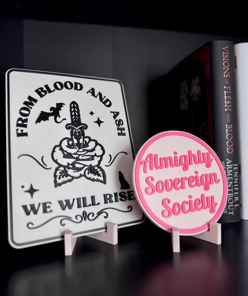 May include: Two white signs with black text and graphics. The sign on the left says "From Blood and Ash We Will Rise" with a black rose, a dagger, a bat, and a wolf. The sign on the right is pink and says "Almighty Sovereign Society".
