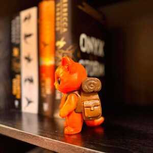 May include: An orange figurine of a cat with a brown backpack stands on a shelf. The figurine is in front of a row of books with the title "Onyx Storm" visible. The cat is facing left.