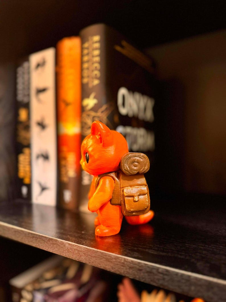 May include: An orange figurine of a cat with a brown backpack stands on a shelf. The figurine is in front of a row of books with the title "Onyx Storm" visible. The cat is facing left.
