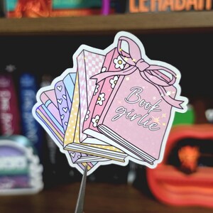May include: A die-cut sticker featuring a stack of five books in pastel colors. The front book is pink with white daisies, a large pink bow, and the text "Book girlie" in cursive. Other books feature patterns including hearts, yellow polka dots, and a pink checkerboard.
