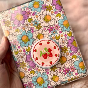 Puede incluir: Una funda de teléfono con estampado floral y un agarre emergente con diseño de fresas. La funda presenta un colorido diseño de flores en tonos azules, morados y blancos. El agarre emergente tiene un fondo rosa con fresas.
