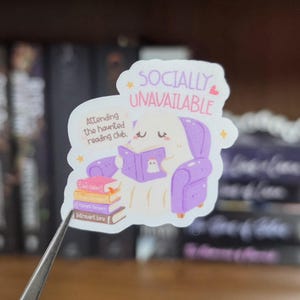 K&ouml;nnte beinhalten: Ein gestanzter Aufkleber mit einem wei&szlig;en Geist, der in einem violetten Sessel sitzt und ein violettes Buch liest. Der Text lautet SOCIALLY UNAVAILABLE und Attending the haunted reading club. Ein Stapel aus vier bunten B&uuml;chern liegt neben dem Geist.