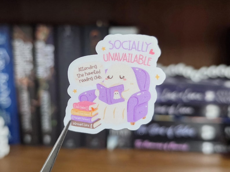 K&ouml;nnte beinhalten: Ein gestanzter Aufkleber mit einem wei&szlig;en Geist, der in einem violetten Sessel sitzt und ein violettes Buch liest. Der Text lautet SOCIALLY UNAVAILABLE und Attending the haunted reading club. Ein Stapel aus vier bunten B&uuml;chern liegt neben dem Geist.