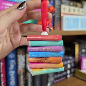 May include: A miniature ornament featuring a stack of seven books by Sarah J. Maas, including A Court of Thorns and Roses, suspended by a red ribbon. The spines are colored red, teal, purple, blue, orange, yellow, and light green.