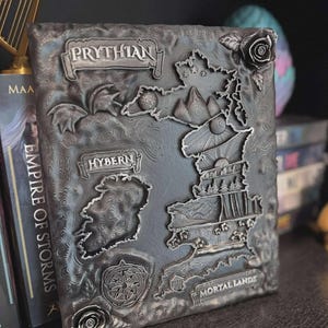 May include: A detailed, metallic gray book cover with a map design. The cover features raised relief elements, including the words "Prythian," "Hybern," and "Mortal Lands." Decorative roses and a compass rose are also present. The book is displayed on a shelf with other books.