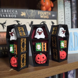 May include: Three black coffin-shaped shelves with gold-colored decorative trim. Each shelf contains a white ghost reading a book, a green and orange book set with a skull emblem, and a small orange jack-o'-lantern at the bottom.