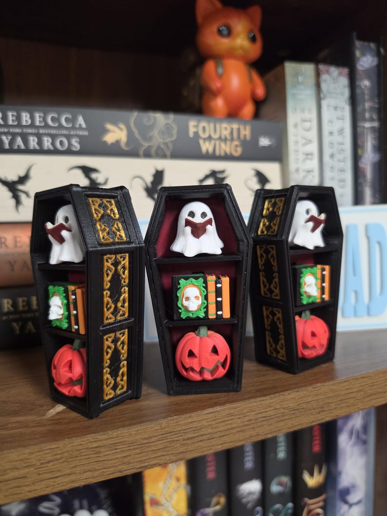 May include: Three black coffin-shaped shelves with gold-colored decorative trim. Each shelf contains a white ghost reading a book, a green and orange book set with a skull emblem, and a small orange jack-o'-lantern at the bottom.