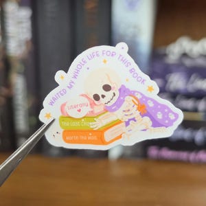 K&ouml;nnte beinhalten: Ein gestanzter Aufkleber mit einem Cartoon-Skelett, das einen violetten Umhang mit Geistermustern tr&auml;gt und auf einem Stapel aus drei B&uuml;chern liegt. Der Text lautet "WAITED MY WHOLE LIFE FOR THIS BOOK", "Literally", "The Last Chapter" und "Worth The Wait".