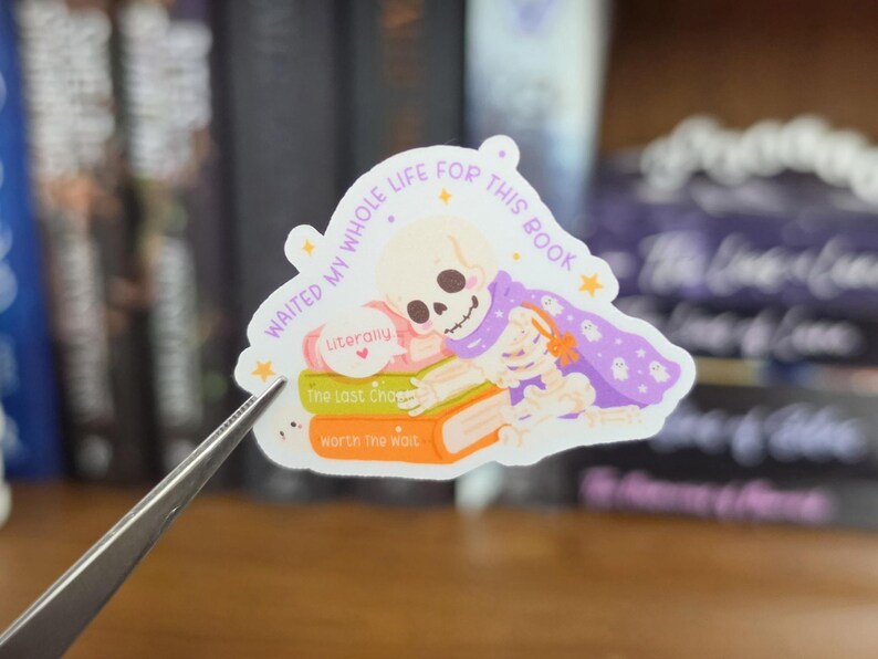 K&ouml;nnte beinhalten: Ein gestanzter Aufkleber mit einem Cartoon-Skelett, das einen violetten Umhang mit Geistermustern tr&auml;gt und auf einem Stapel aus drei B&uuml;chern liegt. Der Text lautet "WAITED MY WHOLE LIFE FOR THIS BOOK", "Literally", "The Last Chapter" und "Worth The Wait".