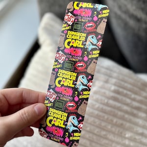 May include: A rectangular bookmark with a black background and colorful graphics. The text "DUNGEON CRAWLER CARL" is prominently displayed, along with images of a dinosaur, donuts, and other elements. The bookmark is held by a hand.