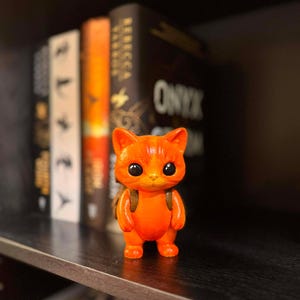May include: A vibrant orange cat figurine with large, dark eyes and a small backpack. The cat is standing on a dark shelf, with books in the background. The figurine is a cute, decorative item.