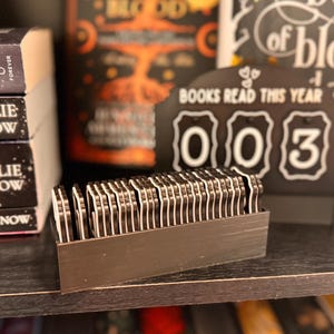 May include: A black wooden shelf displays a stack of books with the author's name "Kylie Snow" and a "Books Read This Year" counter showing "003". A rectangular container holds many small, black and white book-shaped markers.