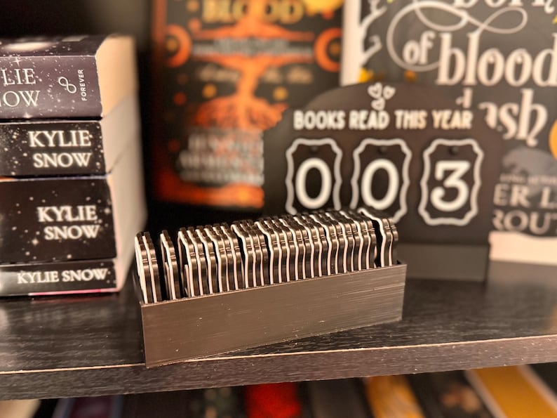 May include: A black wooden shelf displays a stack of books with the author's name "Kylie Snow" and a "Books Read This Year" counter showing "003". A rectangular container holds many small, black and white book-shaped markers.