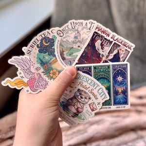 May include: A hand holding a collection of colorful stickers. The stickers feature various designs, including illustrations of a castle, a celestial scene, and text that reads "SURIEL TEA C" and "CRESCENT CITY Postal Service".