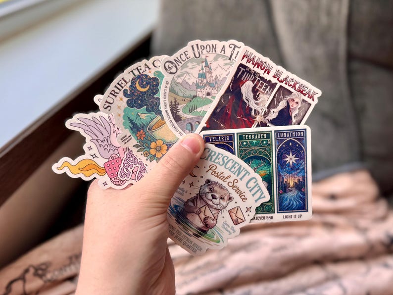 May include: A hand holding a collection of colorful stickers. The stickers feature various designs, including illustrations of a castle, a celestial scene, and text that reads "SURIEL TEA C" and "CRESCENT CITY Postal Service".