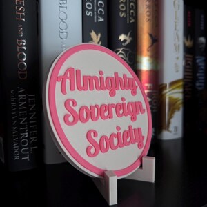 May include: A white circular sign with a pink border and pink text that reads "Almighty Sovereign Society" on a shelf with books.