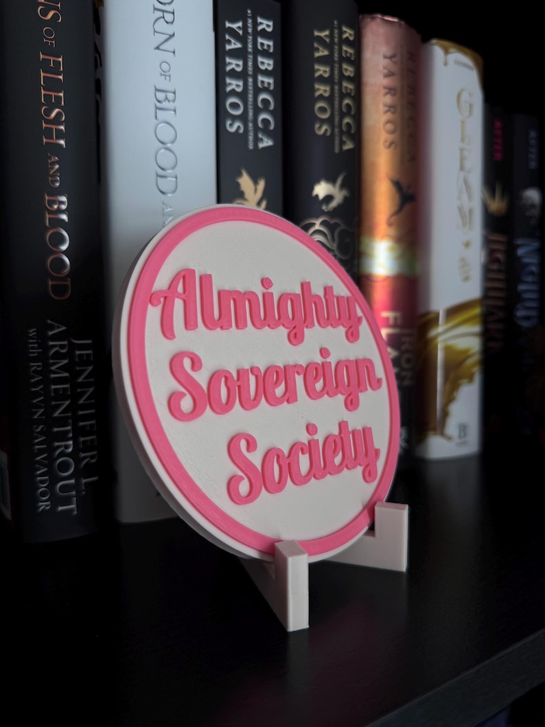 May include: A white circular sign with a pink border and pink text that reads "Almighty Sovereign Society" on a shelf with books.