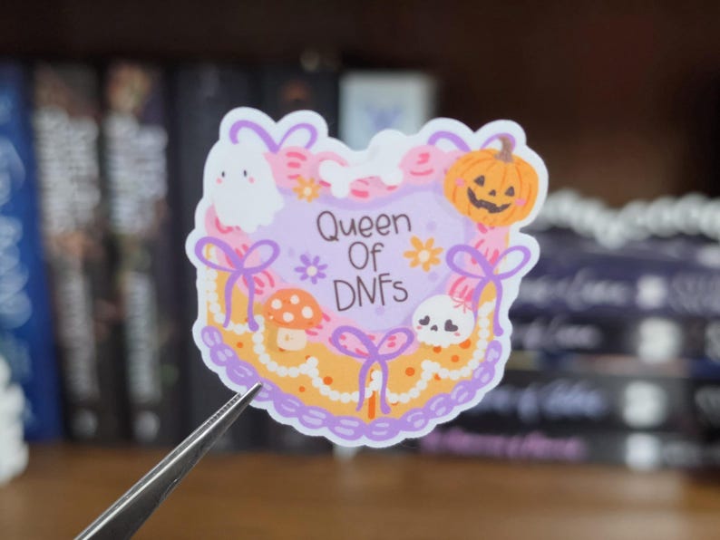 K&ouml;nnte beinhalten: Ein gestanzter Aufkleber mit herzf&ouml;rmigem Design, hellviolettem Zentrum und dem Text Queen Of DNFs. Der Rand ist mit einem wei&szlig;en Geist, einem orangefarbenen K&uuml;rbis, einem kleinen Totenkopf, einem Pilz und violetten Schleifen verziert.